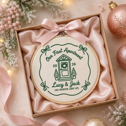 May include: A round wooden embroidery hoop containing cream fabric with green stitched text and graphics. The design features a house, the text "Our First Apartment", "2026", "Lucy &amp; Jack", and "123 NORTH ANY ST.", topped with a light pink satin bow.
