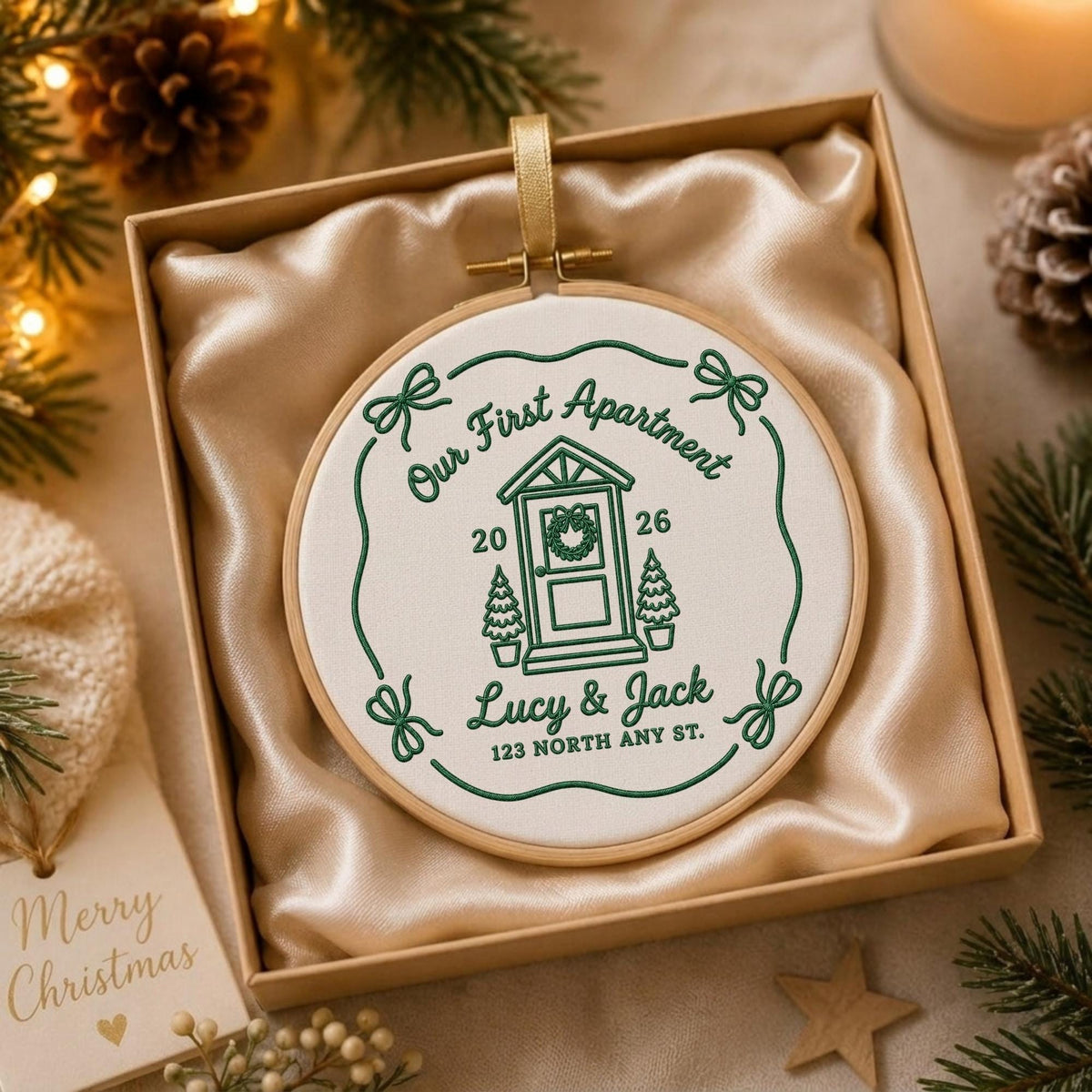 May include: A circular embroidery hoop ornament featuring white fabric with dark green stitched text and graphics. The design includes a house door, two small trees, the text Our First Apartment, 2026, Lucy &amp; Jack, and 123 North Any St.