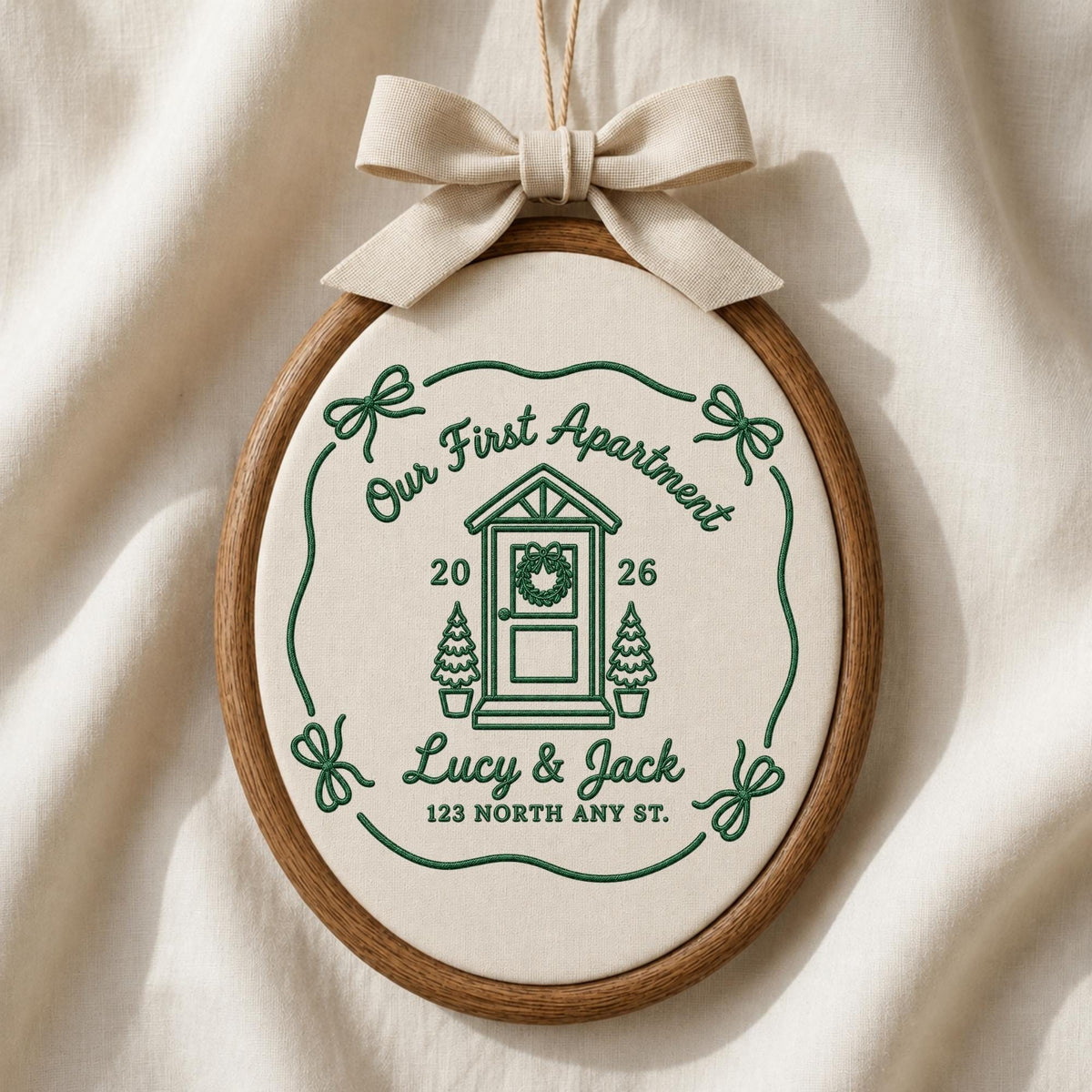 May include: An oval embroidery hoop containing off-white fabric with green stitched text and graphics. The design features a house door, two small trees, the text Our First Apartment, 2026, Lucy &amp; Jack, 123 North Any St., and a beige bow.