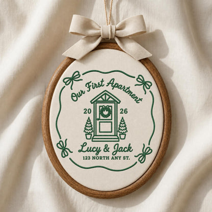 May include: An oval embroidery hoop containing off-white fabric with green stitched text and graphics. The design features a house door, two small trees, the text Our First Apartment, 2026, Lucy &amp; Jack, 123 North Any St., and a beige bow.