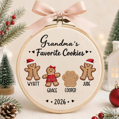 May include: A round wooden embroidery hoop containing cream fabric with embroidered gingerbread figures and a paw print. The text reads Grandma's Favorite Cookies, Wyatt, Grace, Cooper, Jude, and 2026. A light pink satin ribbon bow is tied at the top.