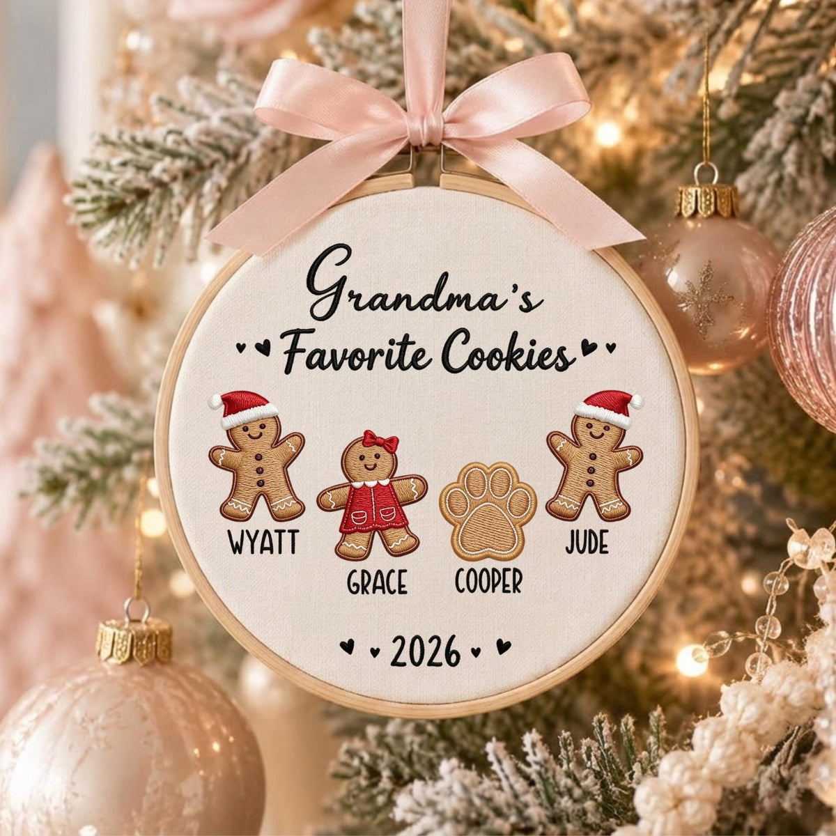 May include: A round wooden embroidery hoop ornament featuring cream fabric with the text Grandma's Favorite Cookies. It includes embroidered gingerbread figures named Wyatt, Grace, and Jude, a paw print labeled Cooper, and the year 2026, topped with a light pink satin ribbon bow.
