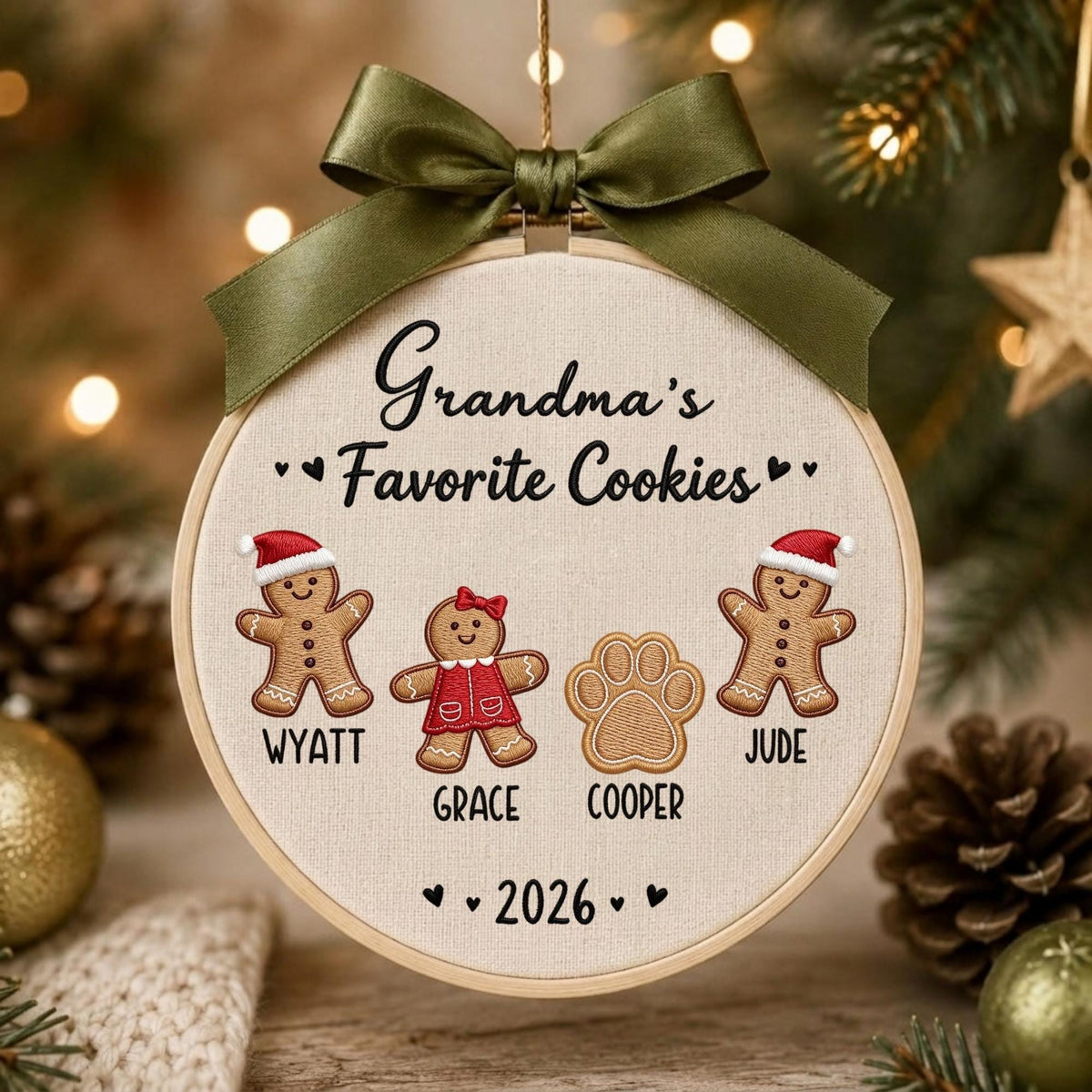 May include: A round embroidery hoop ornament featuring a dark olive green satin ribbon bow at the top. The fabric center displays the embroidered text Grandma's Favorite Cookies, two gingerbread figures, a paw print, and the year 2026.