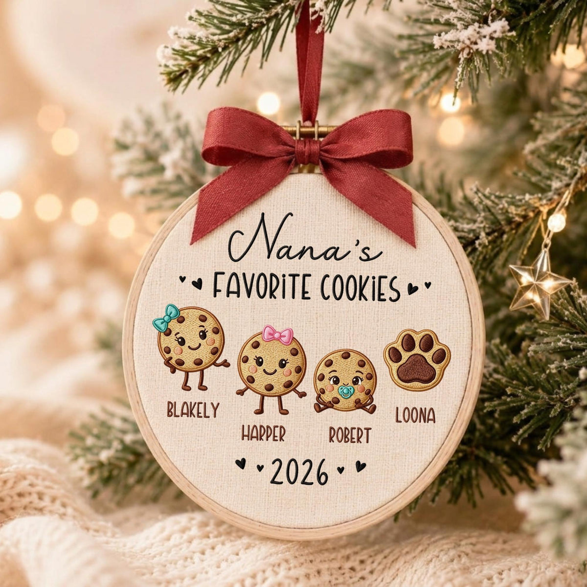May include: A round embroidery hoop ornament featuring the text Nana's FAVORITE COOKIES and 2026. It displays three illustrated cookies with faces and names Blakely, Harper, and Robert, plus a paw print labeled LOONA, topped with a dark red ribbon.