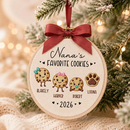 May include: A round embroidery hoop ornament featuring the text Nana's FAVORITE COOKIES and 2026. It displays three illustrated cookies with faces and names Blakely, Harper, and Robert, plus a paw print labeled LOONA, topped with a dark red ribbon.