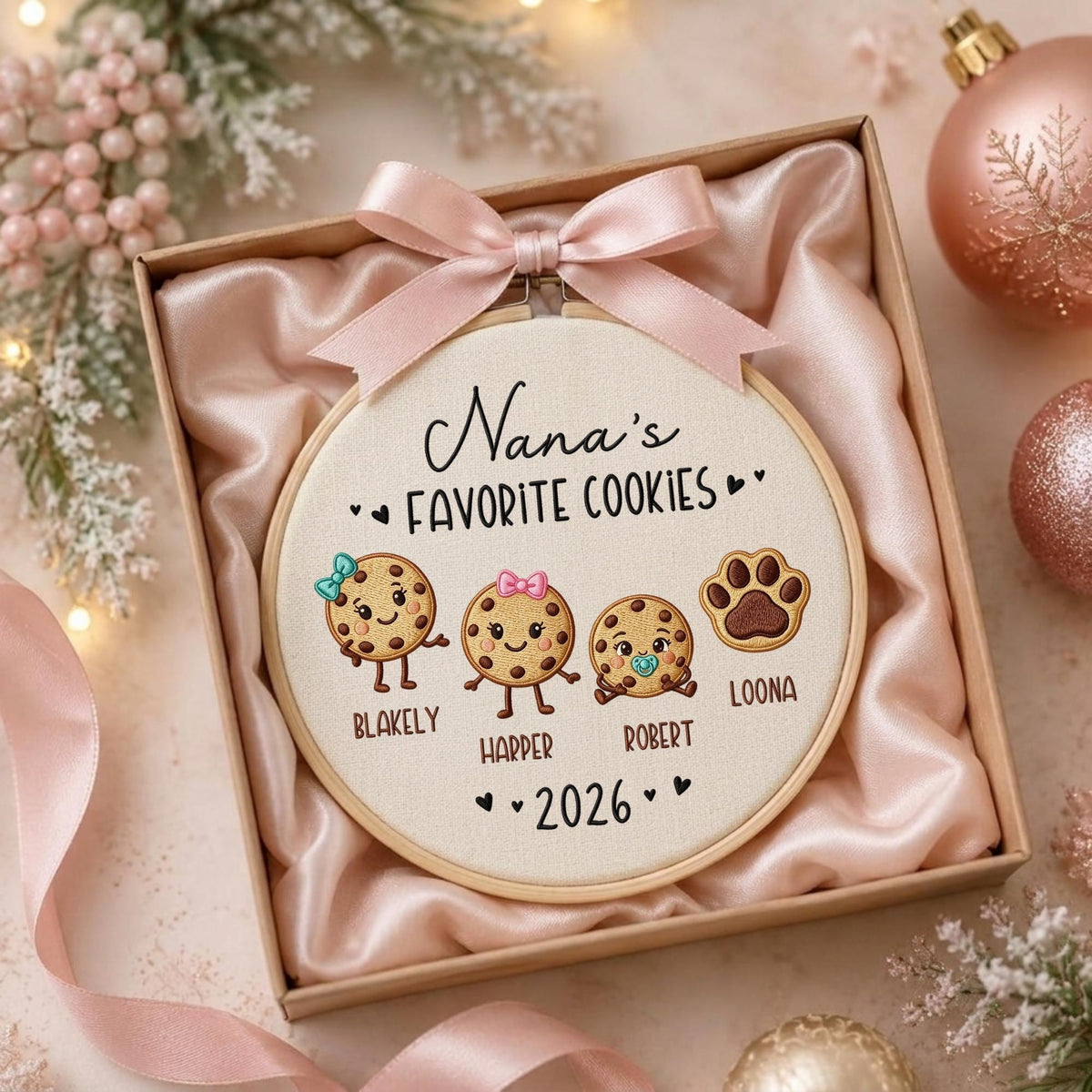 May include: A round embroidery hoop featuring the text Nana's FAVORITE COOKIES and 2026. The design includes three cartoon cookies with faces and a paw print, labeled with the names BLAKELY, HARPER, ROBERT, and LOONA, topped with a pink ribbon.