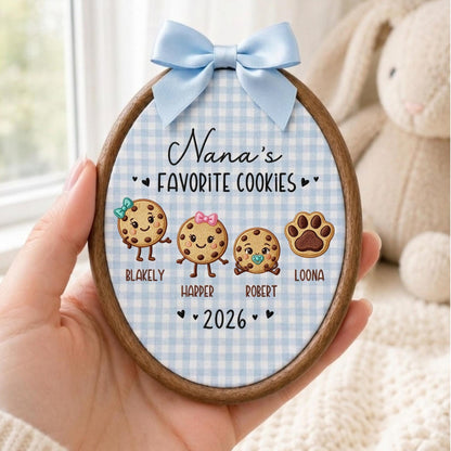 May include: An oval wooden embroidery hoop featuring a light blue gingham fabric background. The text reads Nana's FAVORITE COOKIES, followed by three illustrated cookies with faces and names Blakely, Harper, Robert, a paw print labeled Loona, and 2026.