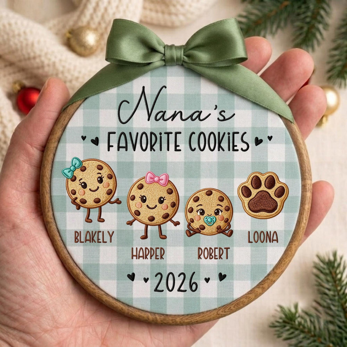 May include: A round wooden embroidery hoop ornament featuring a light green and white buffalo check fabric background. It displays the text Nana's FAVORITE COOKIES, four cartoon cookie characters with names Blakely, Harper, Robert, and Loona, and 2026.