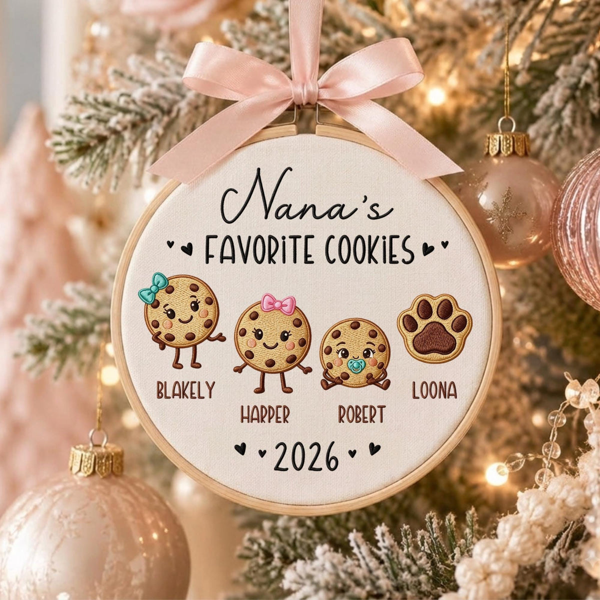 May include: A round wooden embroidery hoop ornament featuring a light pink satin ribbon bow at the top. The fabric center displays the text Nana's FAVORITE COOKIES, three illustrated cookie characters with names BLAKELY, HARPER, ROBERT, a paw print labeled LOONA, and the year 2026.