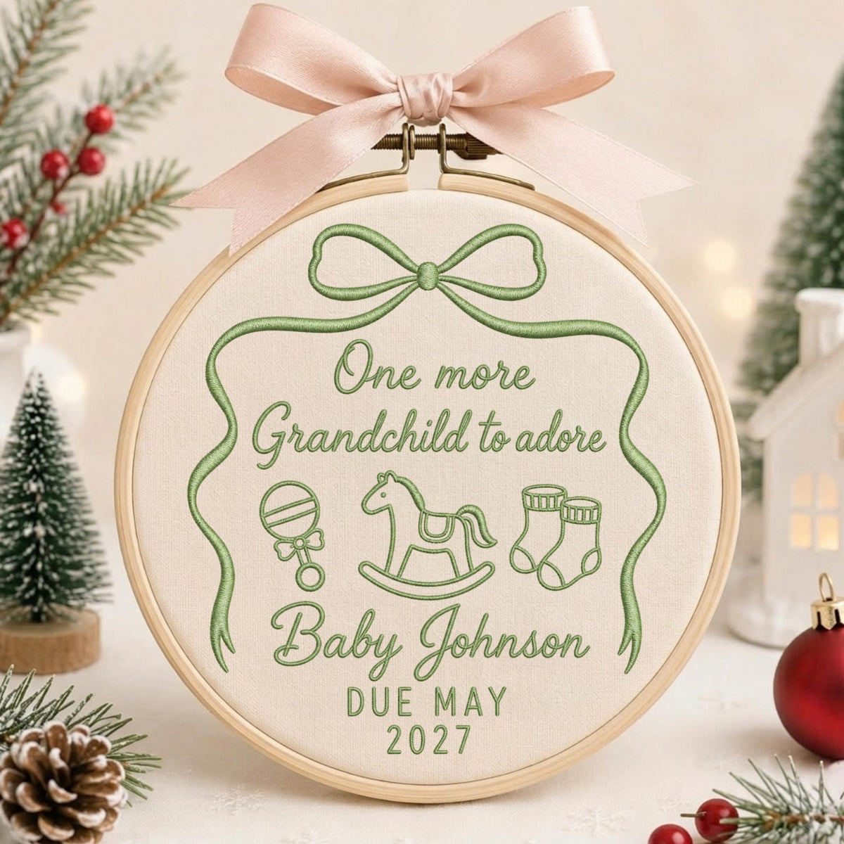 May include: A round wooden embroidery hoop containing cream-colored fabric with green embroidered text reading One more Grandchild to adore, Baby Johnson, DUE MAY 2027, and icons of a rattle, rocking horse, and booties, topped with a pale pink ribbon.