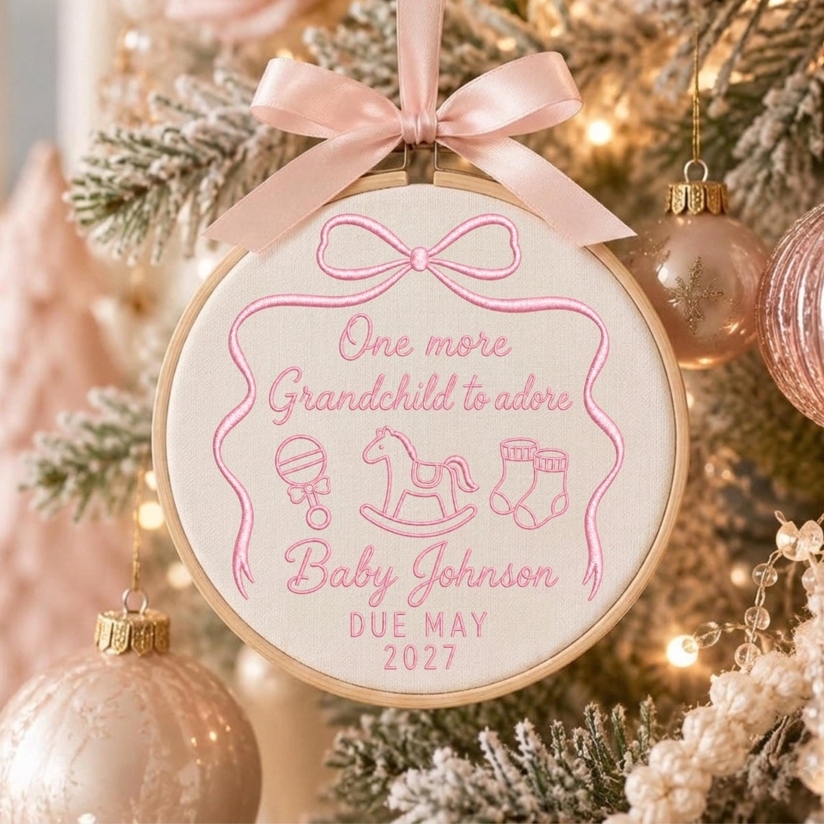 May include: A round embroidery hoop ornament featuring light pink stitching on cream fabric. The text reads One more Grandchild to adore, Baby Johnson, and DUE MAY 2027, with embroidered icons of a rattle, rocking horse, and baby booties.