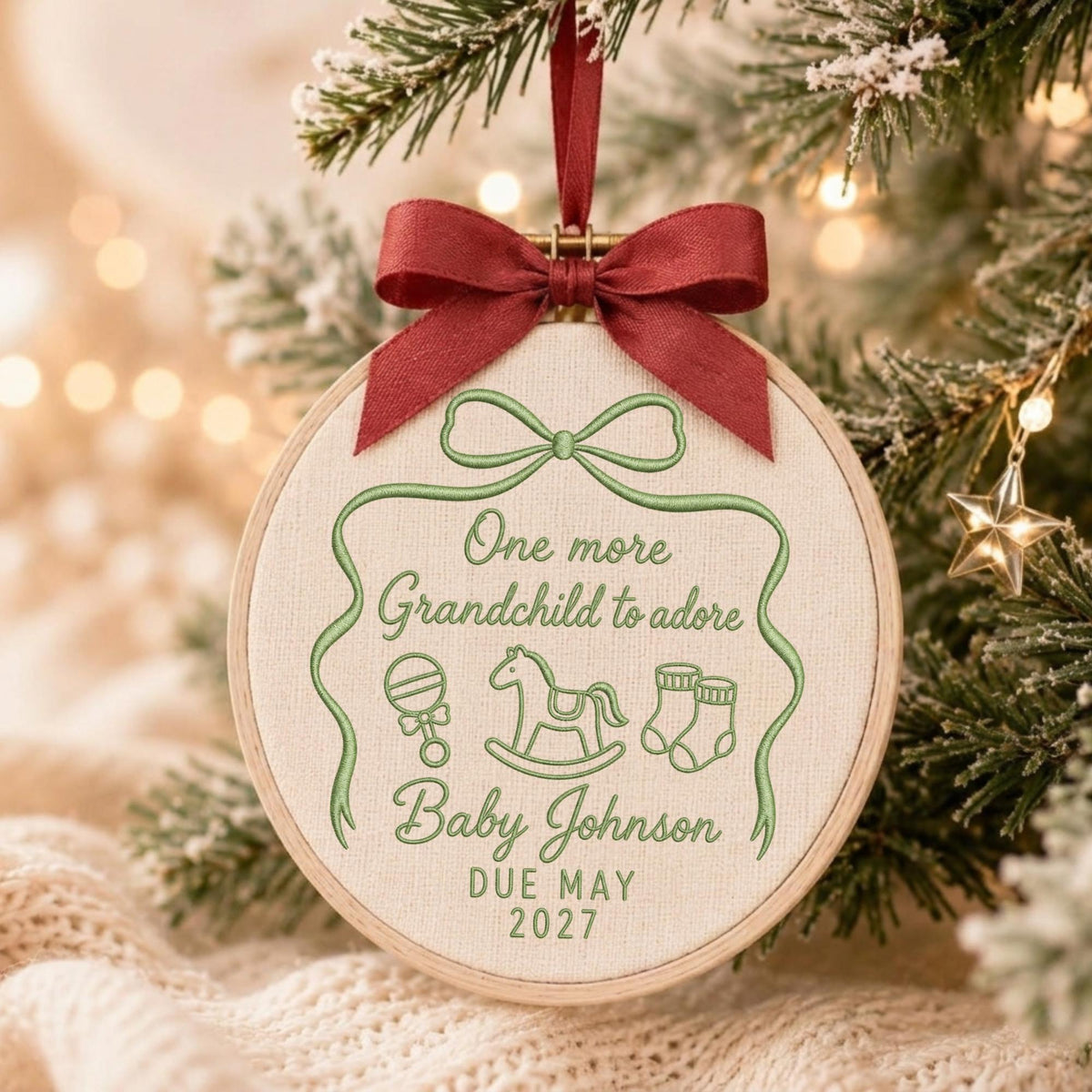 May include: A round embroidery hoop ornament featuring cream fabric with green embroidery. The text reads One more Grandchild to adore, Baby Johnson, DUE MAY 2027. It includes icons of a rattle, rocking horse, and booties, topped with a dark red ribbon.