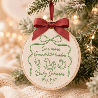 May include: A round embroidery hoop ornament featuring cream fabric with green embroidery. The text reads One more Grandchild to adore, Baby Johnson, DUE MAY 2027. It includes icons of a rattle, rocking horse, and booties, topped with a dark red ribbon.