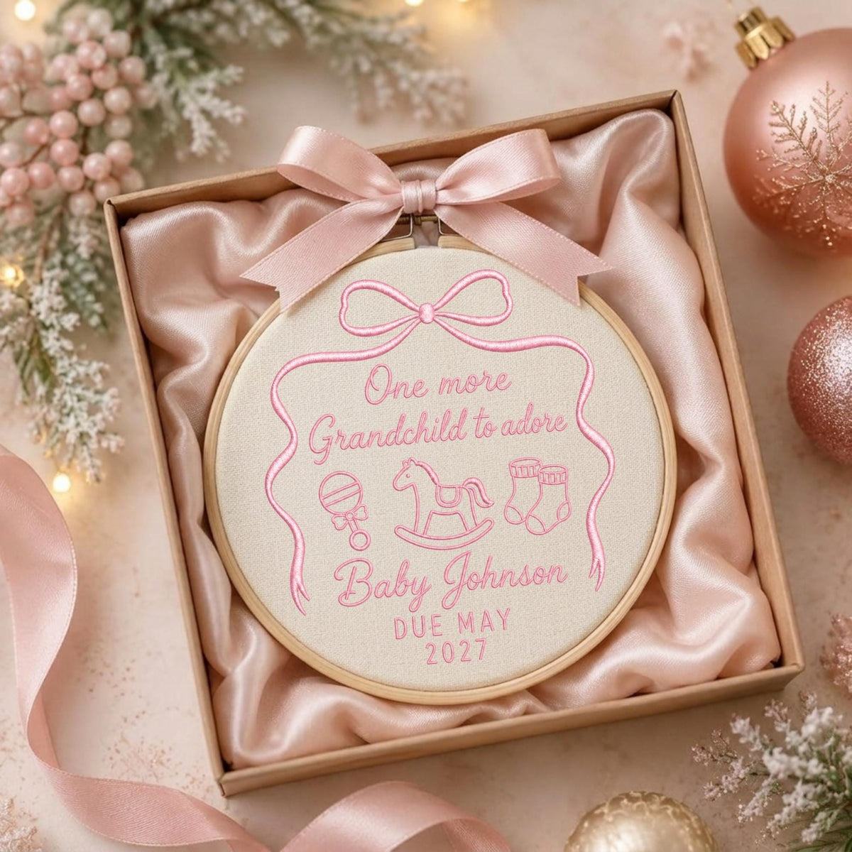 May include: A round wooden embroidery hoop containing cream fabric with pink stitched text reading One more Grandchild to adore, Baby Johnson, and DUE MAY 2027, featuring icons of a rattle, rocking horse, and booties, topped with a pink satin bow.