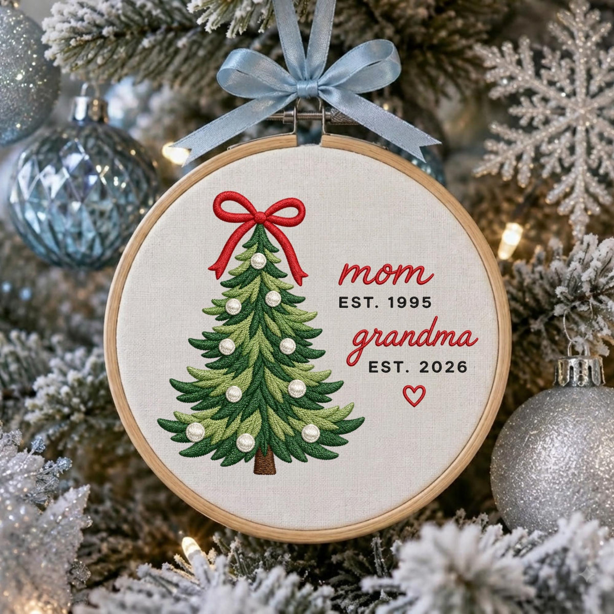 May include: A circular wooden embroidery hoop ornament featuring a stitched green Christmas tree with a red bow on top and white pearl-like beads. The text reads mom EST. 1995, grandma EST. 2026, and a small red heart. A light blue ribbon hangs from the top.