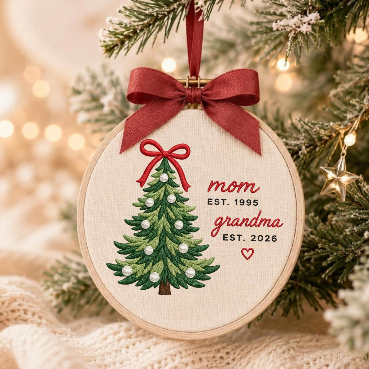 May include: A round wooden embroidery hoop ornament featuring a stitched green Christmas tree with white pearl accents and a red bow topper. The text reads mom EST. 1995 and grandma EST. 2026 with a small red heart. A dark red ribbon hangs from the top.