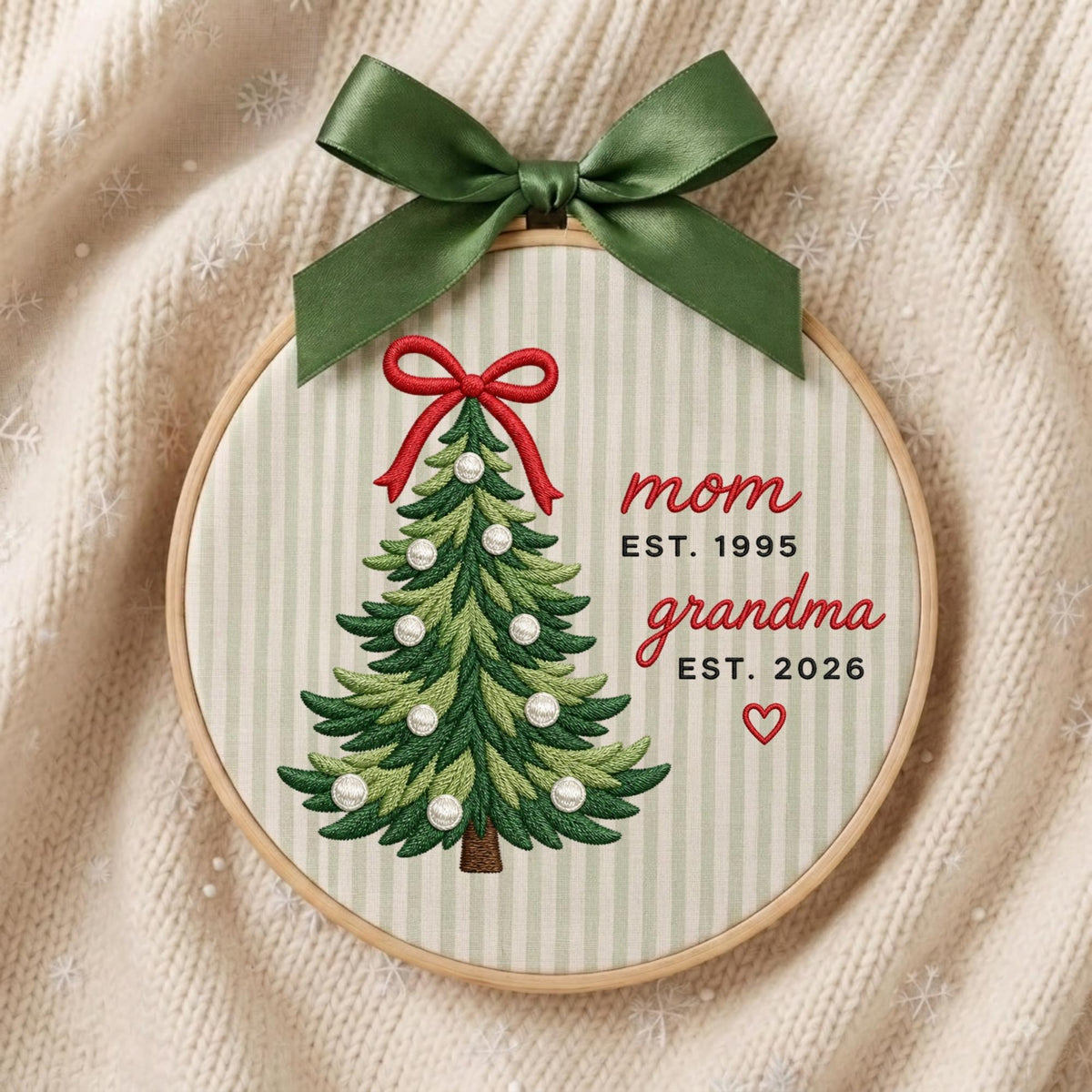 May include: A circular embroidery hoop featuring a light green and white striped fabric background. An embroidered green Christmas tree with white pearl accents and a red bow sits beside the text mom EST. 1995 and grandma EST. 2026 with a heart.