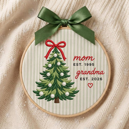 May include: A circular embroidery hoop featuring a light green and white striped fabric background. An embroidered green Christmas tree with white pearl accents and a red bow sits beside the text mom EST. 1995 and grandma EST. 2026 with a heart.