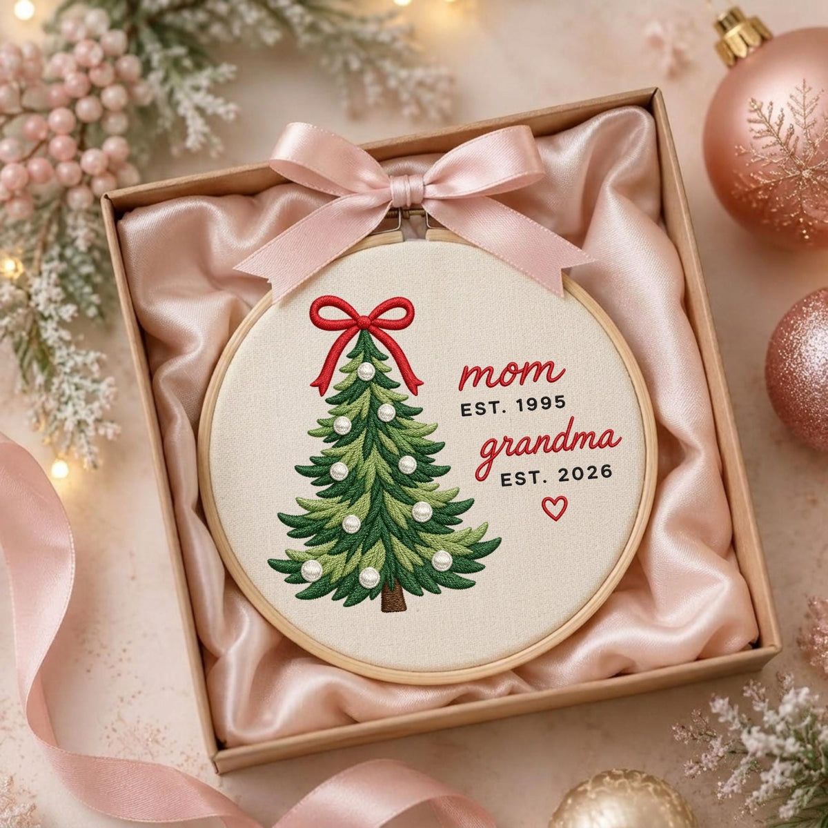 May include: A round wooden embroidery hoop containing a fabric panel with a stitched green Christmas tree decorated with white pearls and a red bow. The text reads mom EST. 1995 and grandma EST. 2026 with a small red heart. A pink satin ribbon is tied at the top.