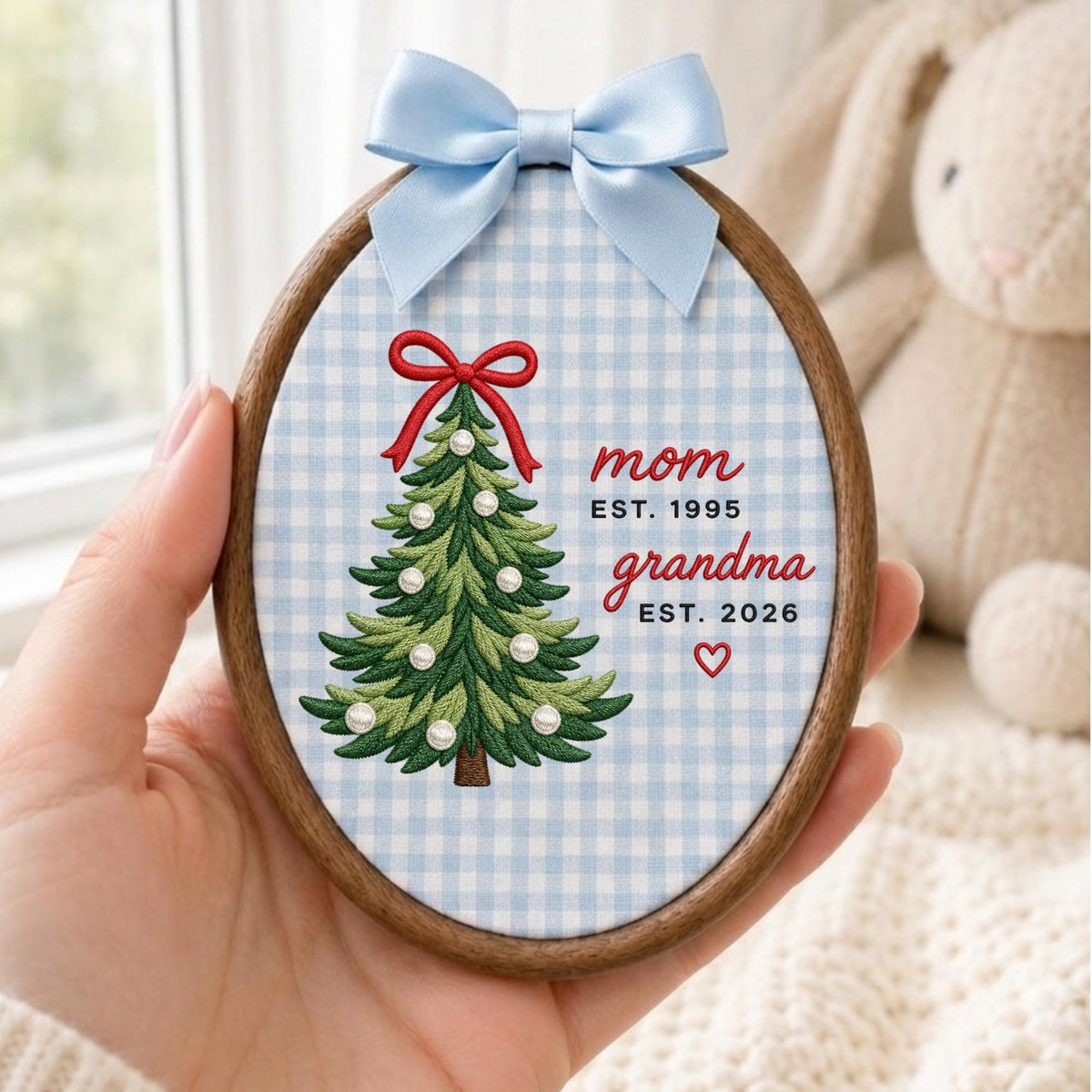 May include: An oval wooden embroidery hoop containing fabric with a light blue and white gingham pattern. It features an embroidered green Christmas tree with a red bow and white pearl accents, plus the text mom EST. 1995, grandma EST. 2026, and a small red heart. A light blue satin ribbon is tied at the top.