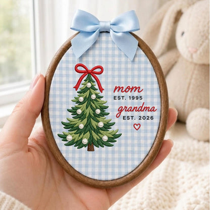 May include: An oval wooden embroidery hoop containing fabric with a light blue and white gingham pattern. It features an embroidered green Christmas tree with a red bow and white pearl accents, plus the text mom EST. 1995, grandma EST. 2026, and a small red heart. A light blue satin ribbon is tied at the top.