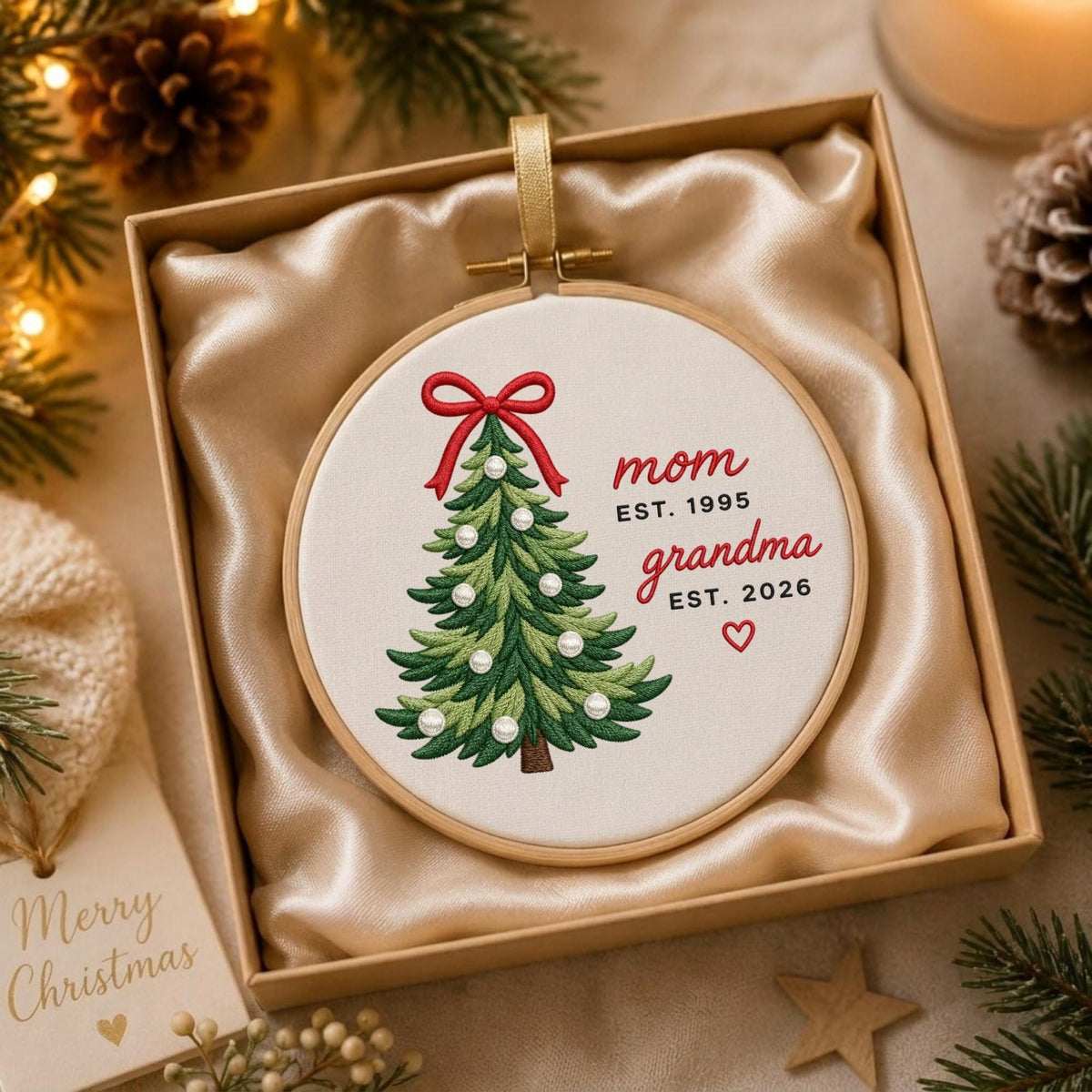 May include: A round embroidery hoop ornament featuring a green Christmas tree with a red bow and white pearl accents. The text reads: mom EST. 1995, grandma EST. 2026, with a small red heart. The ornament is presented in a square box with satin fabric.