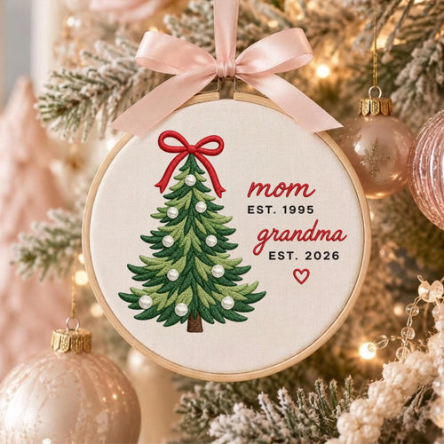 May include: A round wooden embroidery hoop ornament featuring a stitched green Christmas tree with a red bow and white pearl accents. The text reads mom EST. 1995 and grandma EST. 2026 with a small red heart. A light pink satin ribbon hangs from the top.
