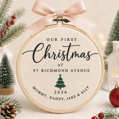 May include: A round wooden embroidery hoop containing cream-colored fabric with black embroidered text reading Our First Christmas at 97 Richmond Avenue, Mummy, Daddy, Jake &amp; Ally, and the year 2026, featuring a green embroidered Christmas tree and a pink bow.