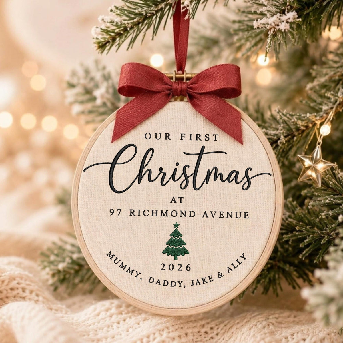 May include: A round embroidery hoop ornament featuring cream-colored fabric with black text reading Our First Christmas at 97 Richmond Avenue, a green embroidered pine tree, and the text 2026 Mummy, Daddy, Jake &amp; Ally, topped with a red ribbon bow.