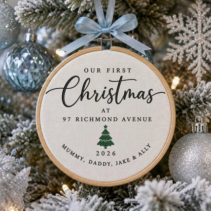 May include: A round wooden embroidery hoop ornament featuring off-white fabric with black text reading OUR FIRST Christmas AT 97 RICHMOND AVENUE, a green embroidered pine tree, the year 2026, and the names MUMMY, DADDY, JAKE &amp; ALLY.