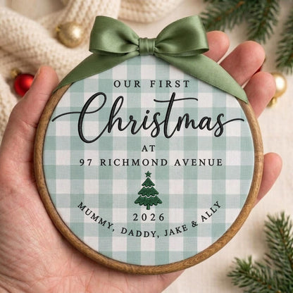 May include: A round wooden embroidery hoop ornament featuring a light green and white buffalo check fabric. The text reads OUR FIRST Christmas AT 97 RICHMOND AVENUE, with a green tree icon, the year 2026, and MUMMY, DADDY, JAKE &amp; ALLY. A sage green satin ribbon bow is tied at the top.