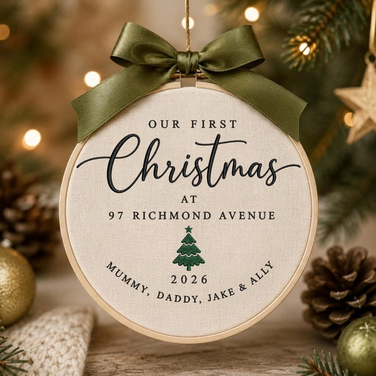 May include: A round embroidery hoop ornament featuring cream-colored fabric with black embroidered text that reads Our First Christmas at 97 Richmond Avenue, Mummy, Daddy, Jake &amp; Ally, and 2026, with a green tree icon and an olive green satin bow.