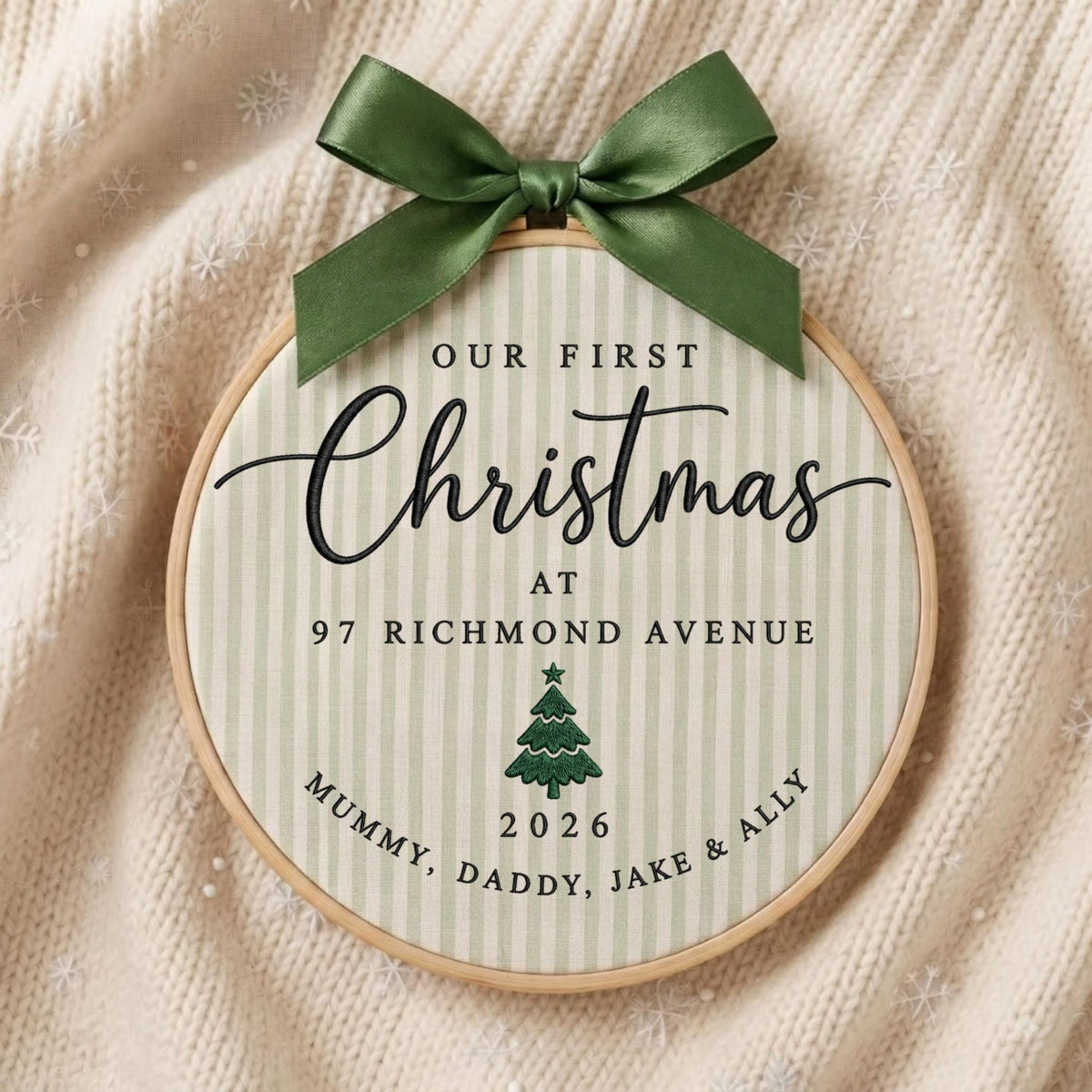 May include: A round wooden embroidery hoop containing fabric with light green vertical stripes. The text reads Our First Christmas at 97 Richmond Avenue, with a green tree icon, the year 2026, and the names Mummy, Daddy, Jake &amp; Ally. A dark green satin ribbon bow is tied at the top.