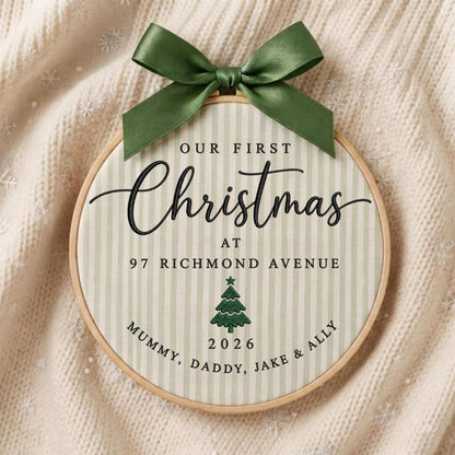 May include: A round wooden embroidery hoop containing fabric with light green vertical stripes. The text reads Our First Christmas at 97 Richmond Avenue, with a green tree icon, the year 2026, and the names Mummy, Daddy, Jake &amp; Ally. A dark green satin ribbon bow is tied at the top.