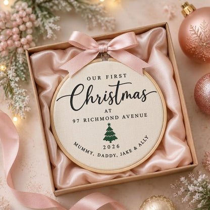 May include: A round wooden embroidery hoop containing cream-colored fabric with black text reading OUR FIRST Christmas AT 97 RICHMOND AVENUE, a green tree icon, and the text 2026 MUMMY, DADDY, JAKE &amp; ALLY. A light pink satin ribbon bow is attached to the top.