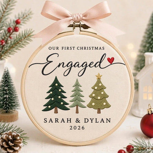 May include: A circular wooden embroidery hoop containing cream-colored fabric with the text OUR FIRST CHRISTMAS Engaged, the names SARAH &amp; DYLAN, and the year 2026. Three embroidered Christmas trees and a small red heart are featured, topped with a pink ribbon.