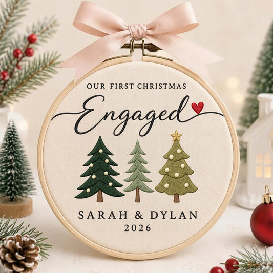 May include: A circular wooden embroidery hoop containing cream-colored fabric with the text OUR FIRST CHRISTMAS Engaged, the names SARAH &amp; DYLAN, and the year 2026. Three embroidered Christmas trees and a small red heart are featured, topped with a pink ribbon.