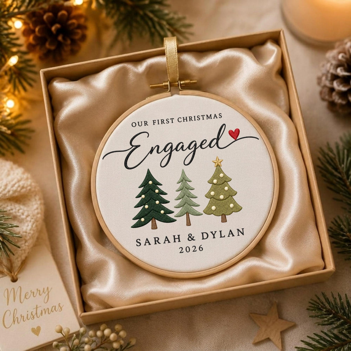 May include: A round embroidery hoop ornament featuring white fabric with the text Our First Christmas Engaged, three embroidered Christmas trees, and the names Sarah &amp; Dylan with the year 2026, all presented in a beige gift box with satin lining.