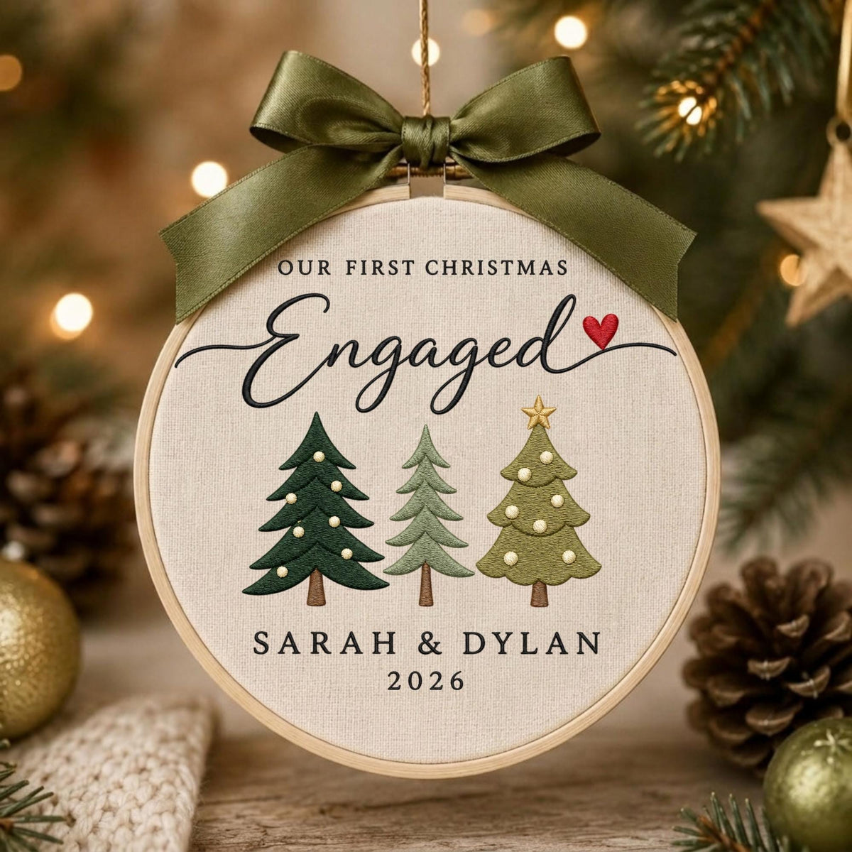 May include: A round wooden embroidery hoop ornament featuring cream fabric with the text OUR FIRST CHRISTMAS ENGAGED and a red heart. Below are three embroidered pine trees and the names SARAH &amp; DYLAN with the year 2026. An olive green satin ribbon bow is tied at the top.