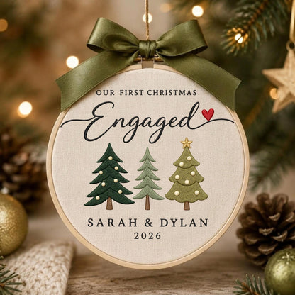 May include: A round wooden embroidery hoop ornament featuring cream fabric with the text OUR FIRST CHRISTMAS ENGAGED and a red heart. Below are three embroidered pine trees and the names SARAH &amp; DYLAN with the year 2026. An olive green satin ribbon bow is tied at the top.