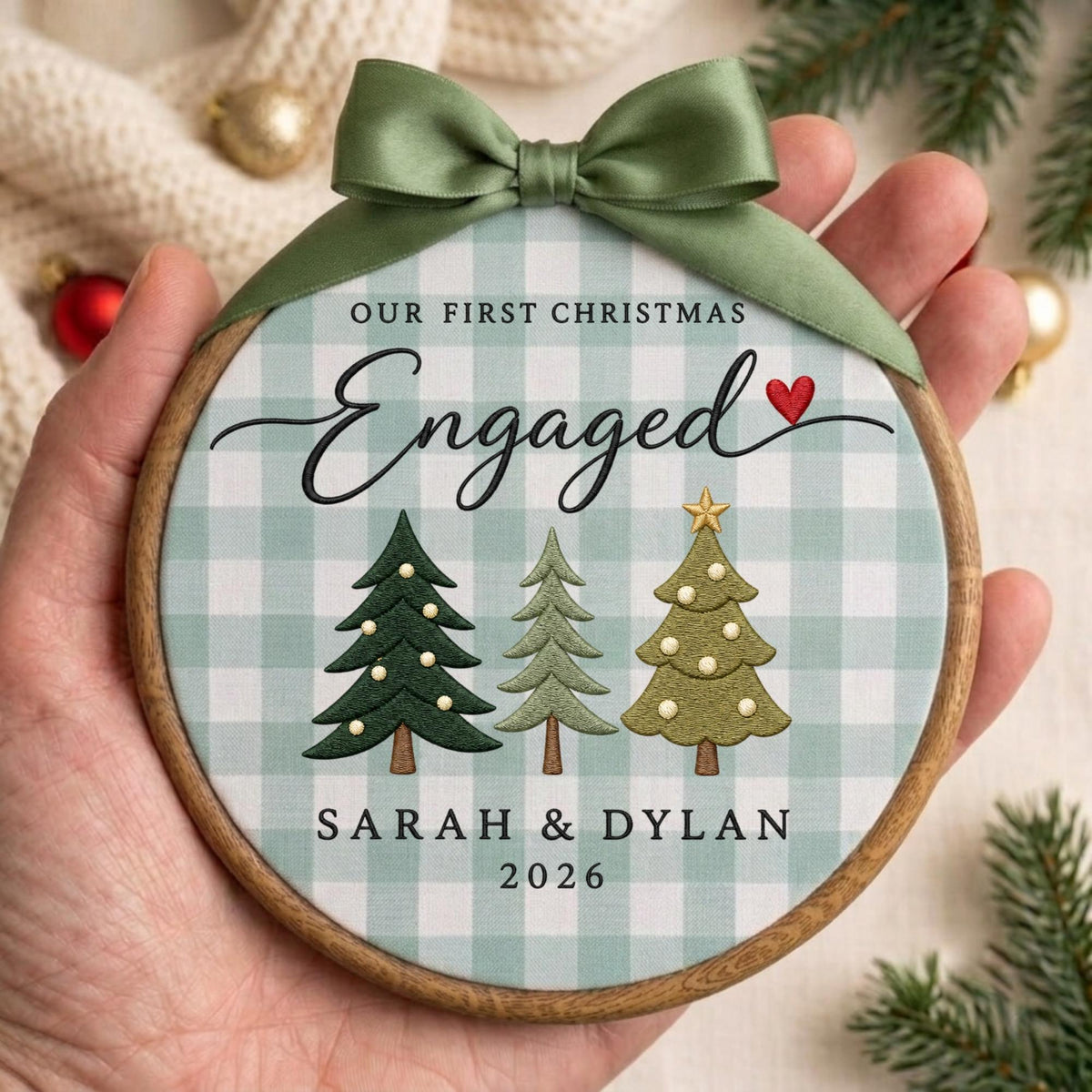 May include: A round wooden embroidery hoop ornament featuring a light green and white buffalo check fabric. The text reads OUR FIRST CHRISTMAS Engaged, SARAH &amp; DYLAN, and 2026. It includes three embroidered Christmas trees and a green satin bow.