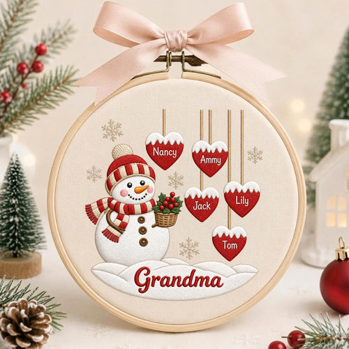 May include: A round wooden embroidery hoop containing cream-colored fabric with a snowman wearing a red and white striped hat and scarf. The snowman holds a basket of holly. Five red hearts hang above with the names Nancy, Ammy, Jack, Lily, and Tom. The word Grandma is embroidered below, topped with a pale pink ribbon.