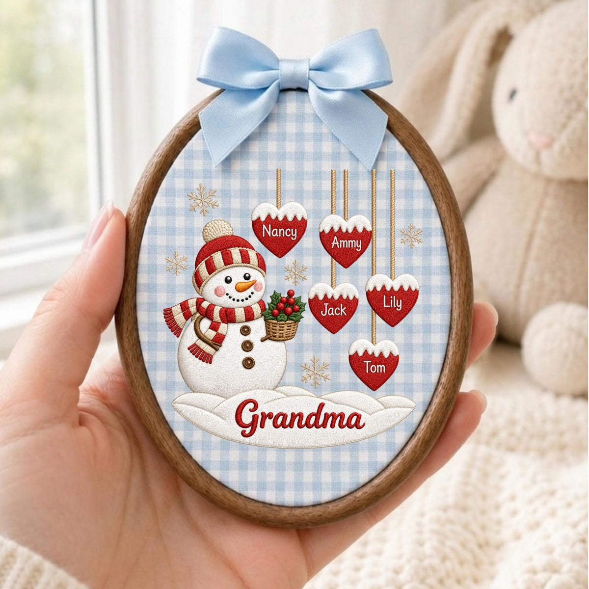 May include: An oval wooden frame containing a fabric print of a snowman wearing a red and white striped hat and scarf, holding a basket. Five red heart ornaments hang above with the names Nancy, Ammy, Jack, Lily, and Tom. The word Grandma is below.