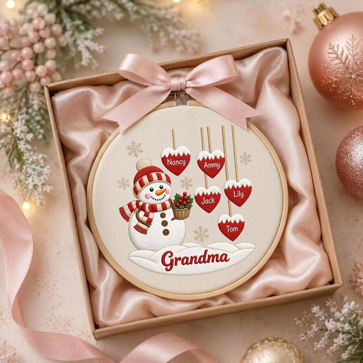 May include: A round wooden embroidery hoop containing a fabric panel with a snowman illustration, red heart ornaments labeled Nancy, Ammy, Jack, Lily, and Tom, and the text Grandma, topped with a pale pink satin ribbon bow inside a gift box.