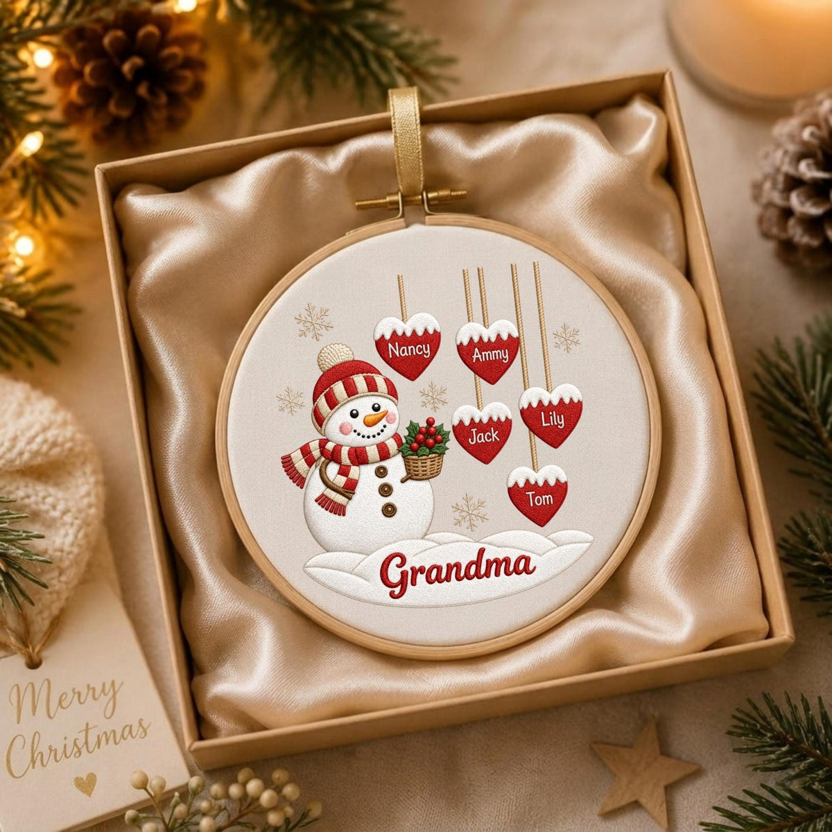 May include: A round embroidery hoop ornament featuring a snowman wearing a red and white striped hat and scarf, holding a basket of holly. Six red heart-shaped ornaments hang above, labeled with the names Nancy, Ammy, Jack, Lily, and Tom, with Grandma below.