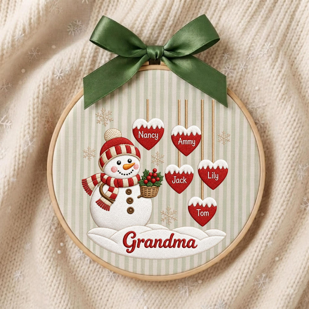 May include: A circular embroidery hoop ornament featuring a snowman wearing a red and white striped hat and scarf, holding a basket of holly. Five red heart-shaped ornaments hang above with the names Nancy, Ammy, Jack, Lily, and Tom. The word Grandma is printed below the snowman, topped with a green ribbon.