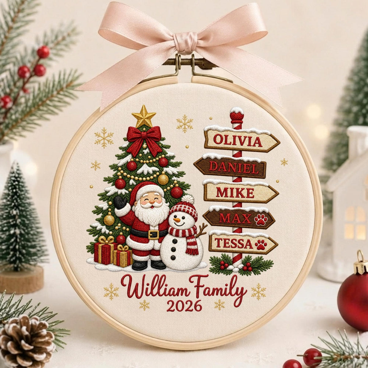 May include: A round wooden embroidery hoop featuring a cream-colored fabric with a Christmas design. It displays a decorated tree, Santa Claus, a snowman, and a signpost with the names Olivia, Daniel, Mike, Max, and Tessa. Text reads William Family 2026, topped with a light pink ribbon bow.