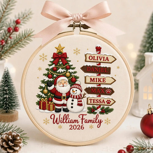 May include: A round wooden embroidery hoop featuring a cream-colored fabric with a Christmas design. It displays a decorated tree, Santa Claus, a snowman, and a signpost with the names Olivia, Daniel, Mike, Max, and Tessa. Text reads William Family 2026, topped with a light pink ribbon bow.
