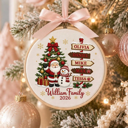 May include: A round embroidery hoop ornament featuring a Christmas tree, Santa Claus, and a snowman. A signpost lists the names Olivia, Daniel, Mike, Max, and Tessa. The text reads William Family 2026. A light pink satin ribbon bow is tied at the top.