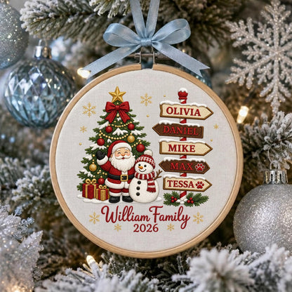 May include: A round wooden embroidery hoop ornament featuring a Christmas tree, Santa Claus, and a snowman. A signpost lists the names Olivia, Daniel, Mike, Max, and Tessa. The text reads William Family 2026. A light blue ribbon is tied at the top.